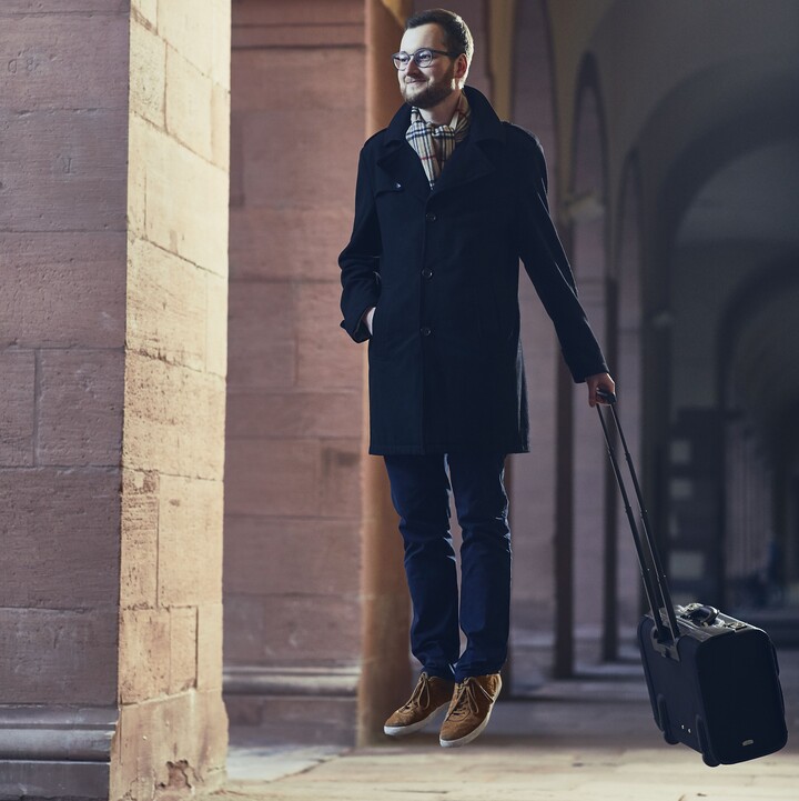 Mann mit Brille, einer dunkelblauen Stoffhose, einem schwarzen langem Mantel und einem gestreiften Schaal steht schwebend über dem Boden mit einem Koffer in der Hand in einem der Gänge vor der Universität.