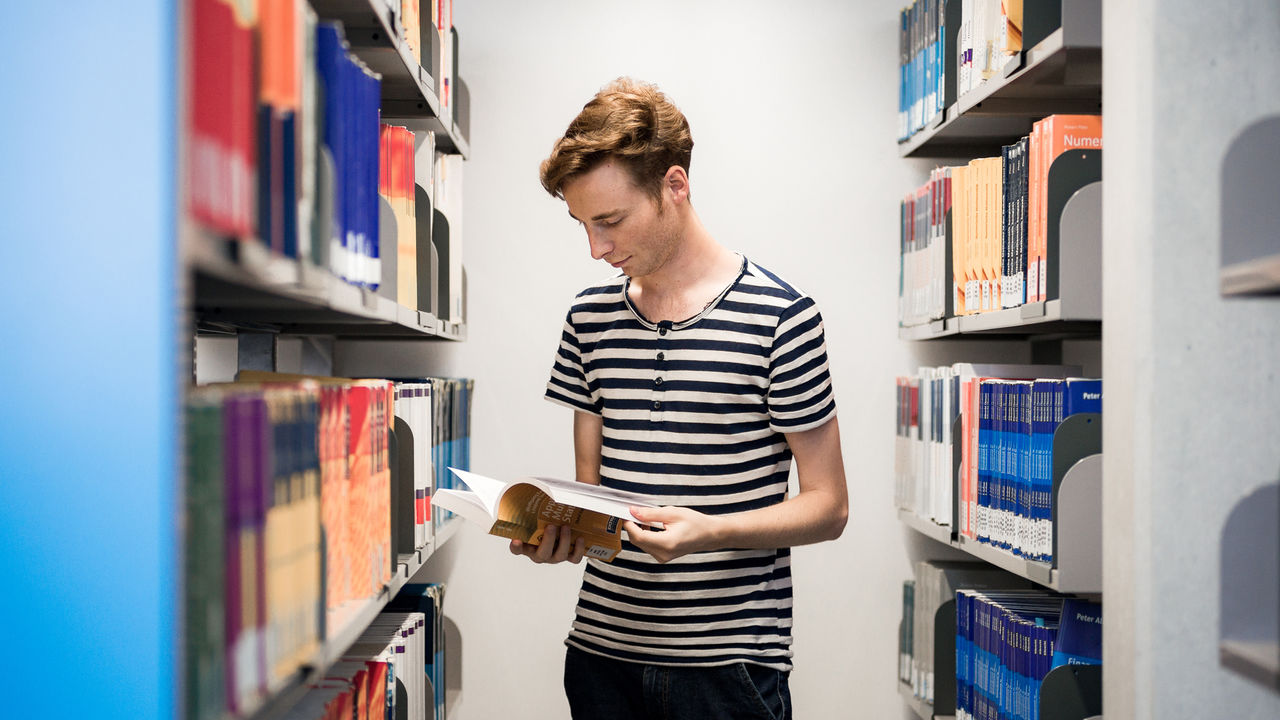 Ein Student hält ein aufgeschlagenes Buch in seinen Händen und blättert darin. Neben ihm stehen gut gefüllte Bücherregale. Der Student trägt kurzes blondes Haar und ein schwarz-weiß gestreiftes T-Shirt.