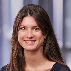 Julia Lier is smiling directly at the camera. She has brown eyes and her long brown hair falls loosely over her shoulders. She is wearing a dark V-neck blouse and a fine golden necklace with a small pendant.