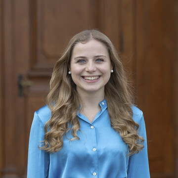 Tonia lacht freundlich in die Kamera. Sie hat lange, dunkelblonde Haare und trägt eine blaue Bluse.