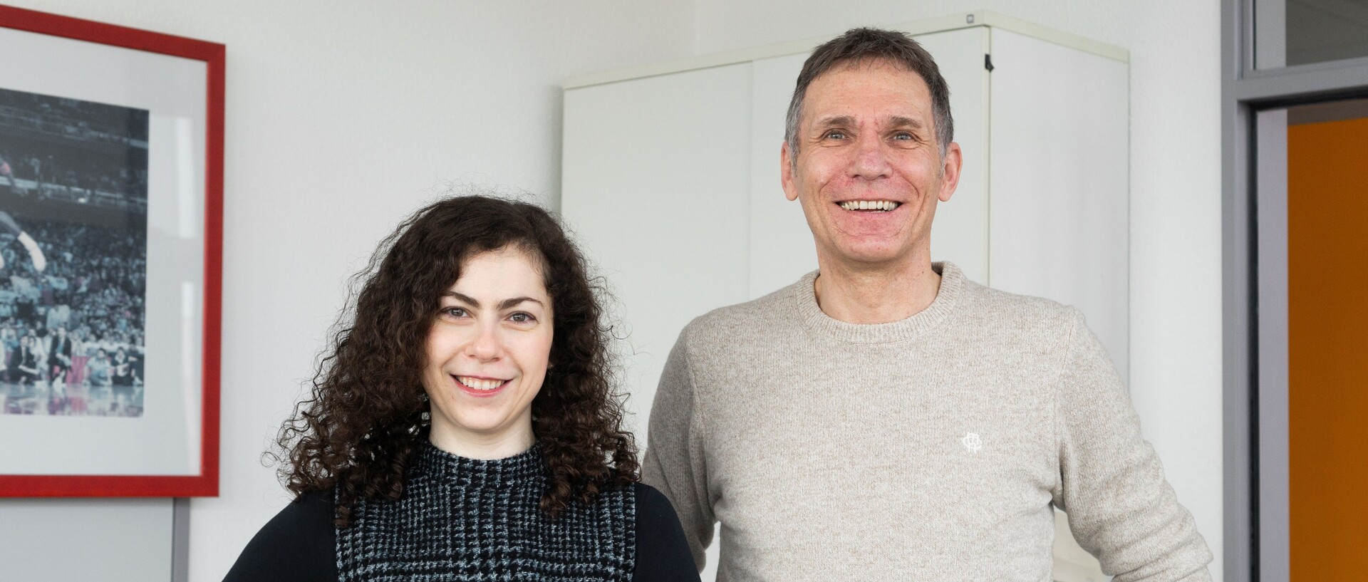 In einem Büro steht Anna Bruk mit schulterlangen schwarzen Locken und einem schwarz-weißen Pollunder über einem schwarzen Shirt links neben Prof. Dr. Herbert Bless in einem beigen Pullover