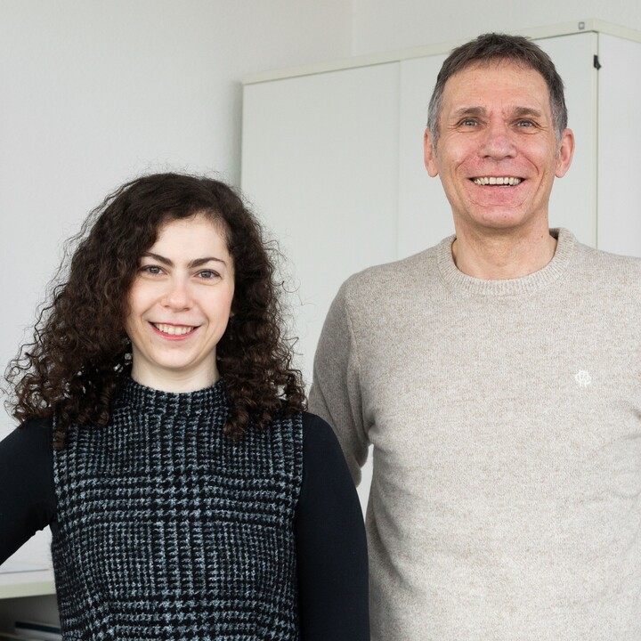 In einem Büro steht Anna Bruk mit schulterlangen schwarzen Locken und einem schwarz-weißen Pollunder über einem schwarzen Shirt links neben Prof. Dr. Herbert Bless in einem beigen Pullover