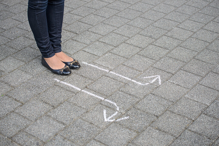 Es sind nur Beine und Füße einer Person zu sehen. Die Person steh auf Pflastersteinboden vor zwei weißen Pfeilen. Ein Pfeil zeigt nach links, der andere nach rechts.