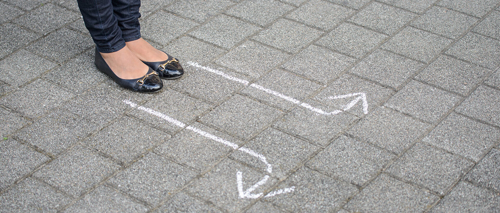 Eine Person steht mit schwarzen Ballerinas und Jeans bekleidet auf grauem Asphaltboden. Vor ihr sind mit Kreide zwei Pfeile auf den Boden gezeichnet, die in entgegengesetze Richtung weisen..