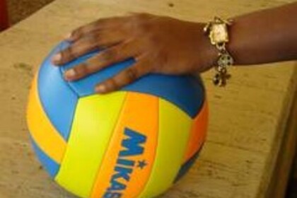 Eine Hand einer schwarzen Person hält einen blau-gelben Volleyball auf dem Tisch fest.