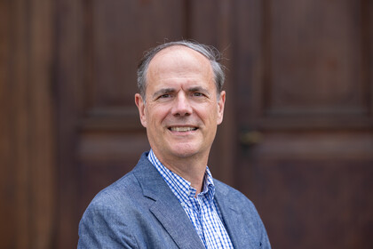 Portrait von Bernhard Ebbinghaus. Er trägt ein blau-weiß karriertes Hemd und einen blauen Blazer und steht lächelnd vor einer braunen Holztüre.