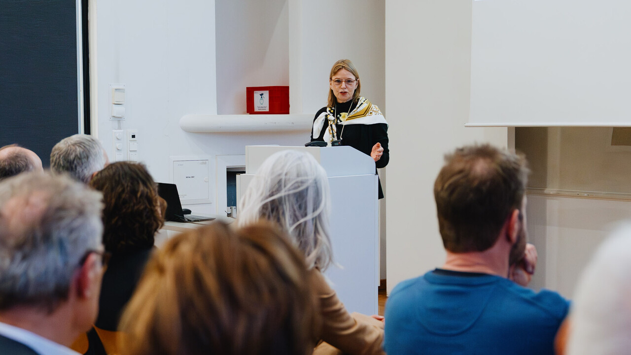 Im Vordergrund sieht man die Hinterkopfe einiger Zuhördenden. Am Rednerpult im Hintergrund steht steht eine Frau in schwarzer Kleidung und Seidenschal. Sie blickt in die Kamera