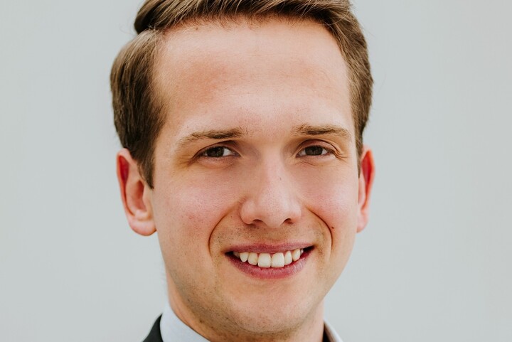 Portrait of  Johannes Gessner. He is wearing a shirt with a black jacket over it and is standing in front of a white background.
