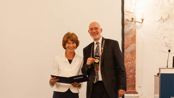Brigitte Fickel trägt einen weißen Blazer und steht mit der offenen schwarzen Mappe neben Prof. Dr. Puhl, der einen schwarzen Anzug trägt und in seiner Hand eine goldene Medaille hält. Im Hintergrund die Marmorwand des Rittersaals der Uni Mannheim.