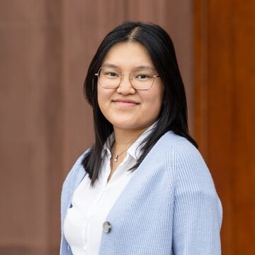 Mädchen mit schwarzen glatten Haaren und Brille. Sie trägt eine weiße Bluse und ein blauen Cardigan