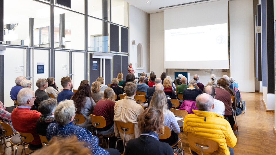 Menschen sitzen in einem Vortragssaal und hören einem Vortrag zu, der auf eine große Leinwand projiziert wird. Eine Person steht vorne und spricht.