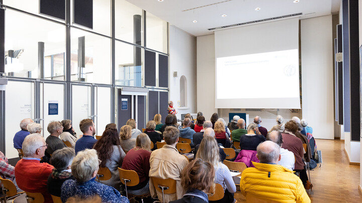 Menschen sitzen in einem Vortragssaal und hören einem Vortrag zu, der auf eine große Leinwand projiziert wird. Eine Person steht vorne und spricht.