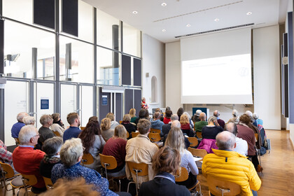 Menschen sitzen in einem Vortragssaal und hören einem Vortrag zu, der auf eine große Leinwand projiziert wird. Eine Person steht vorne und spricht.