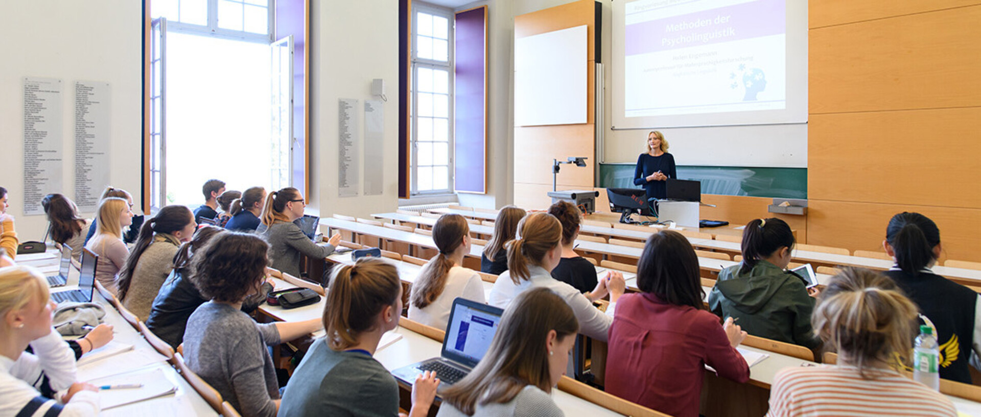 In einem Hörsaal sitzen Studierende, welche auf ihren Laptops tippen. An der Tafel steht eine Frau.