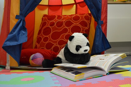 Ein Plüschpanda sitzt in einem farbenfrohen Spielzelt auf bunten Schaumstoff-Puzzlematten. Vor ihm ist ein aufgeschlagenes, illustriertes Buch, in das er scheinbar vertieft liest.
