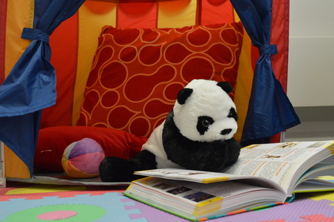 Ein Plüschpanda sitzt in einem farbenfrohen Spielzelt auf bunten Schaumstoff-Puzzlematten. Vor ihm ist ein aufgeschlagenes, illustriertes Buch, in das er scheinbar vertieft liest.