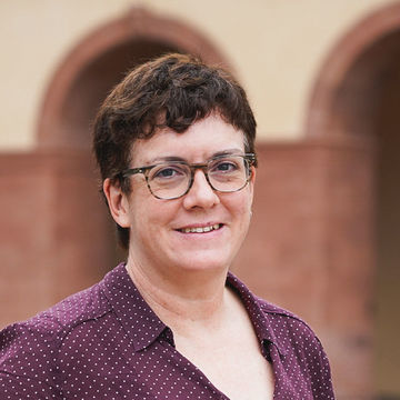 Jane Ossandon-Schönfeld hat braune Haare und eine Kurzhaarfrisur. Sie trägt eine lila Bluse mit weißen Punkten und eine Brille.
