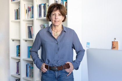 Prof. Wänke trägt eine blaue Bluse und steht vor einem Bücherregal
