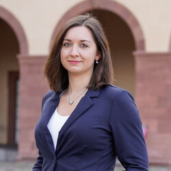 Ein Porträt von Sarina Rottner vor der Fassade des Mannheimer Schlosses. Sie trägt schulterlanges braunes Haar, ein weißes Oberteil und darüber einen dunkelblauen Blazer.