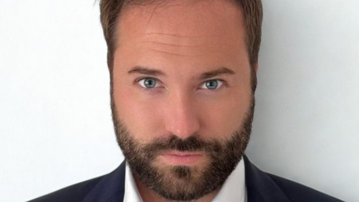 Julian Schenkenhofer hat kurze braune Haare und einen Vollbart. Er trägt ein weißes Hemd und ein dunkelblaues Jackett darüber.