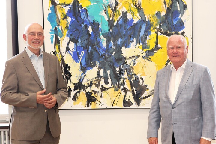 Manfred Fuchs und Thomas Puhl stehen vor einem bunten, abstrakten Gemälde.