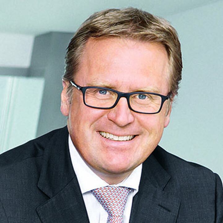 Stefan Fuchs im Porträt mit schwarzer rechteckiger Brille, schwarzem Anzug, weißem Hemd und rot-weißer Krawatte
