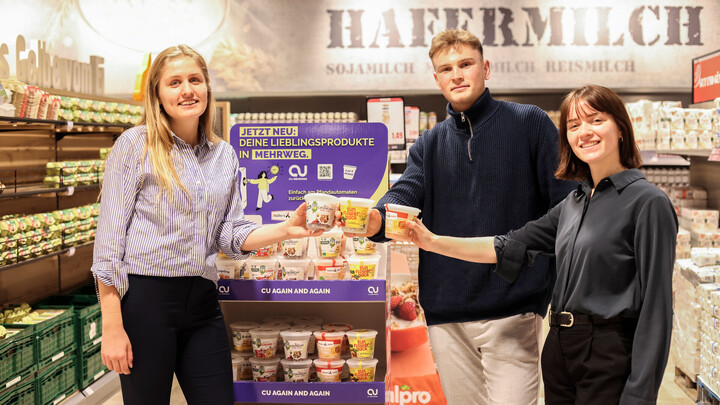 Die Gründer*innen von CU Mehrweg stehen im Supermarkt vor einem Aufsteller und präsentieren ihr Produkt. Alle drei lächeln in die Kamera.