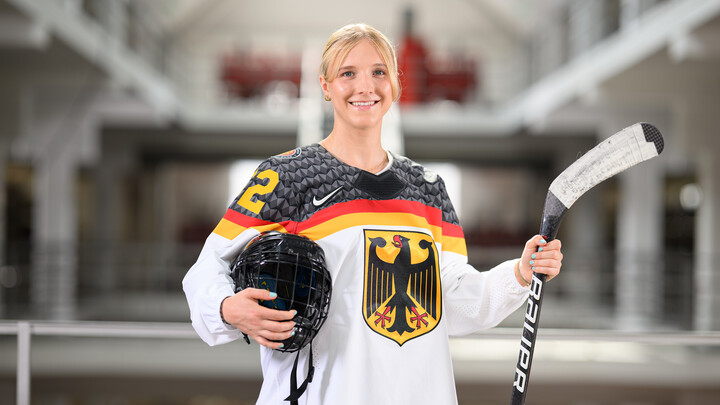 Porträtbild von Lucia Schmitz in der Bibliothek A3. Sie trägt ein Eishockey-Trikot und hält einen Schläger und einen Helm fest.