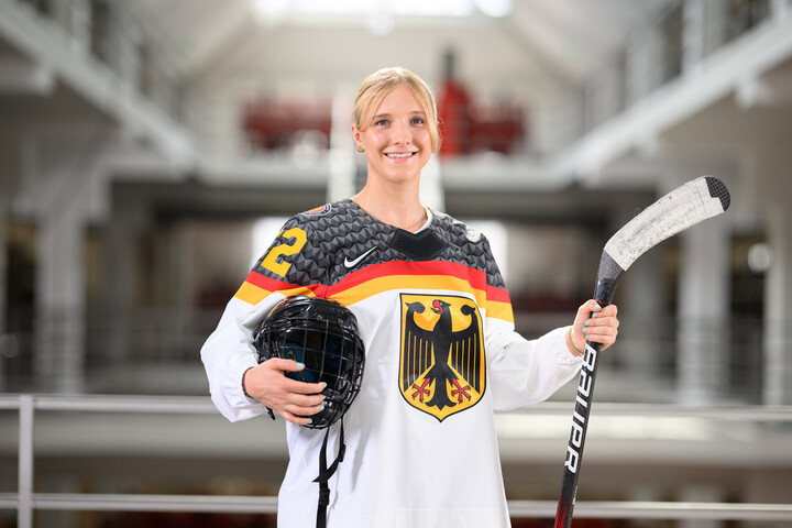 Porträtbild von Lucia Schmitz in der Bibliothek A3. Sie trägt ein Eishockey-Trikot und hält einen Schläger und einen Helm fest.