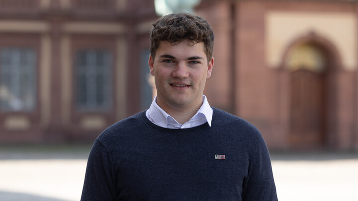 Paul Seidel. He is wearing a blue sweater over a white shirt and is standing on the Ehrenhof in front of one of the statues.
