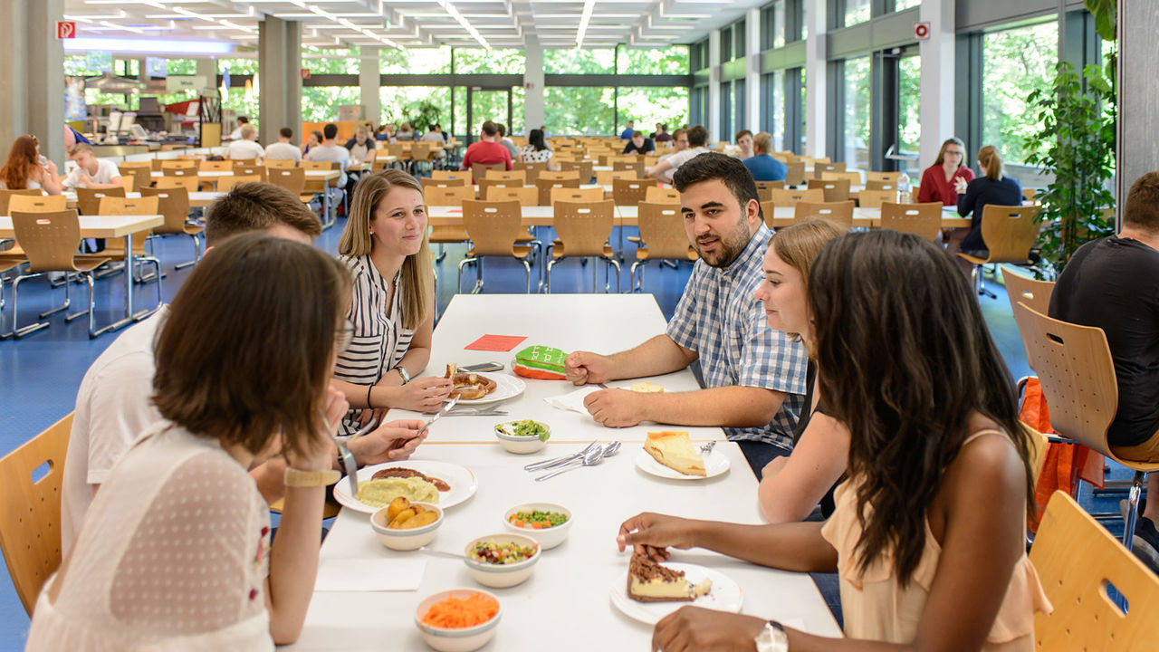 Studierende sitzen gemeinsam in der Mensa an einem Tisch und essen zu Mittag.