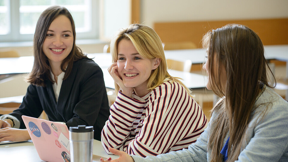 Drei Studentinnen sitzen in einem Seminarraum und lachen. Vor ihnen steht ein aufgeklappter Laptop. Die beiden äußeren Frauen schauen beide zur mittleren.