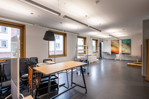 Das Foto zeigt einen großen Büroraum. Er hat vier quadratische Fenster. In ihm sind Möbel zusammengestellt. Man sieht Werkzeuge und ein fahrbares Gerüst. An der kurzen Wand im Hienrgrund sind zwei abstrakte, farbige Bilder aufgehängt.