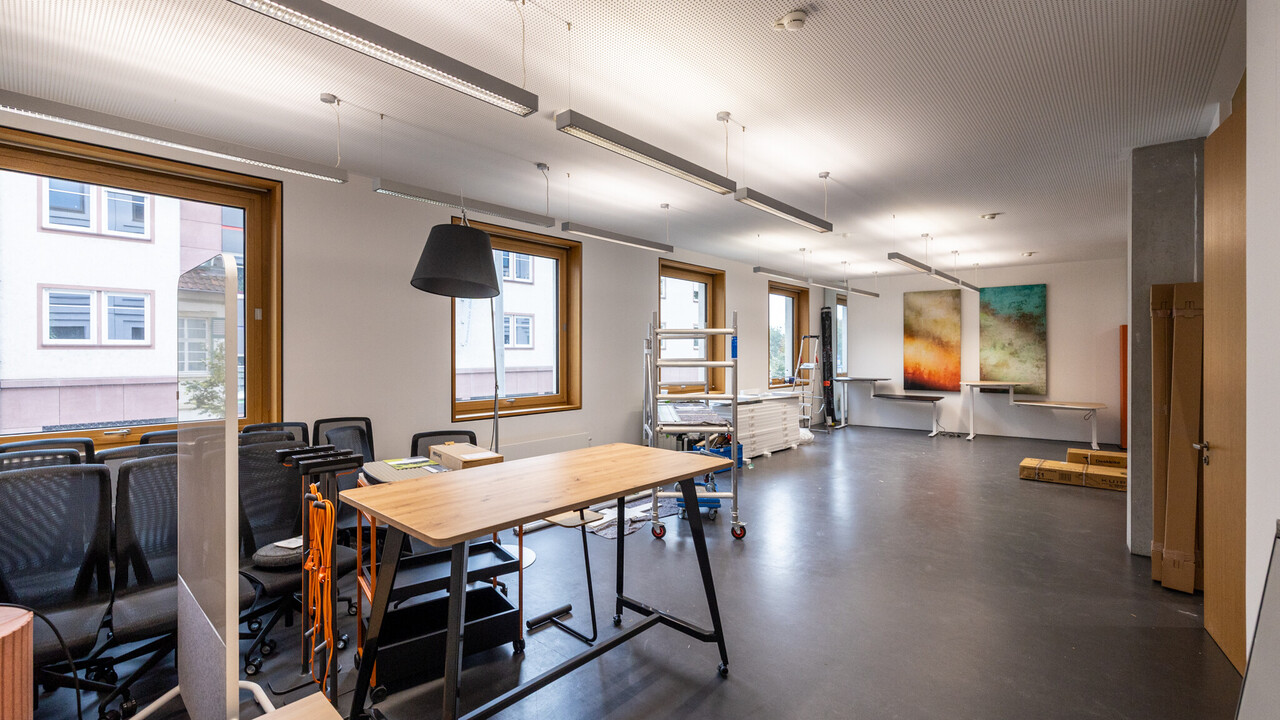 Das Foto zeigt einen großen Büroraum. Er hat vier quadratische Fenster. In ihm sind Möbel zusammengestellt. Man sieht Werkzeuge und ein fahrbares Gerüst. An der kurzen Wand im Hienrgrund sind zwei abstrakte, farbige Bilder aufgehängt.
