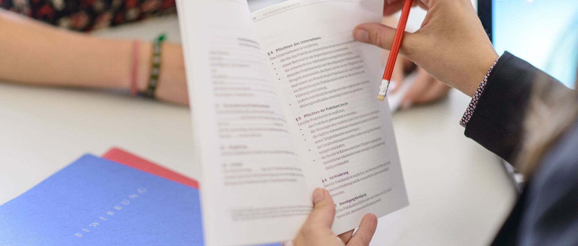 Eine Person hält ein aufgeschlagenes Gesetzbuch in beiden Händen. Darunter liegt eine blaue Mappe. Auf dieser Mappe steht das Wort Bewerbung.