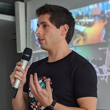 Miguel Arduz has short dark hair. He is wearing a black T-shirt with a colorful print and has a microphone in his hand.