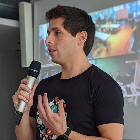 Miguel Arduz has short dark hair. He is wearing a black T-shirt with a colorful print and has a microphone in his hand.