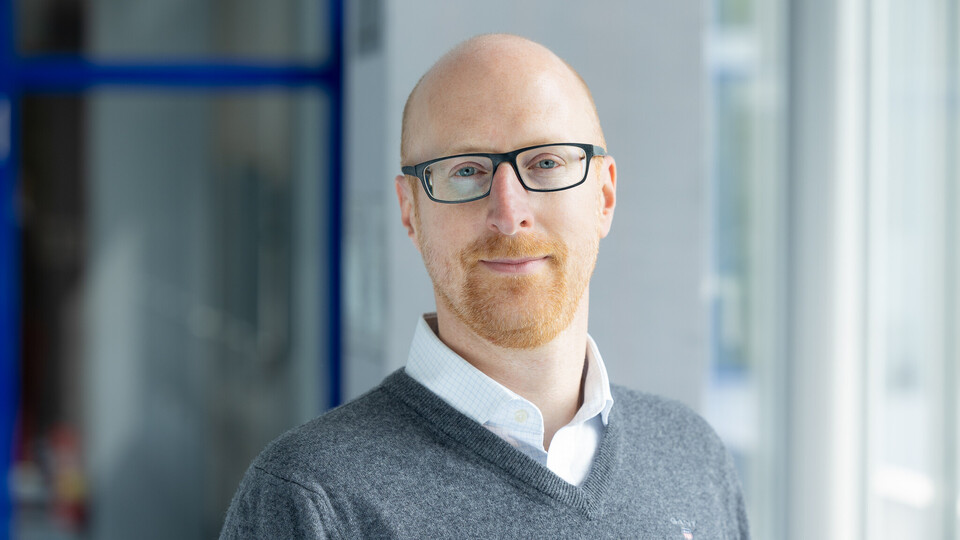 Florian Keusch trägt ein weißes Hemd und darüber einen grauen Pullover.