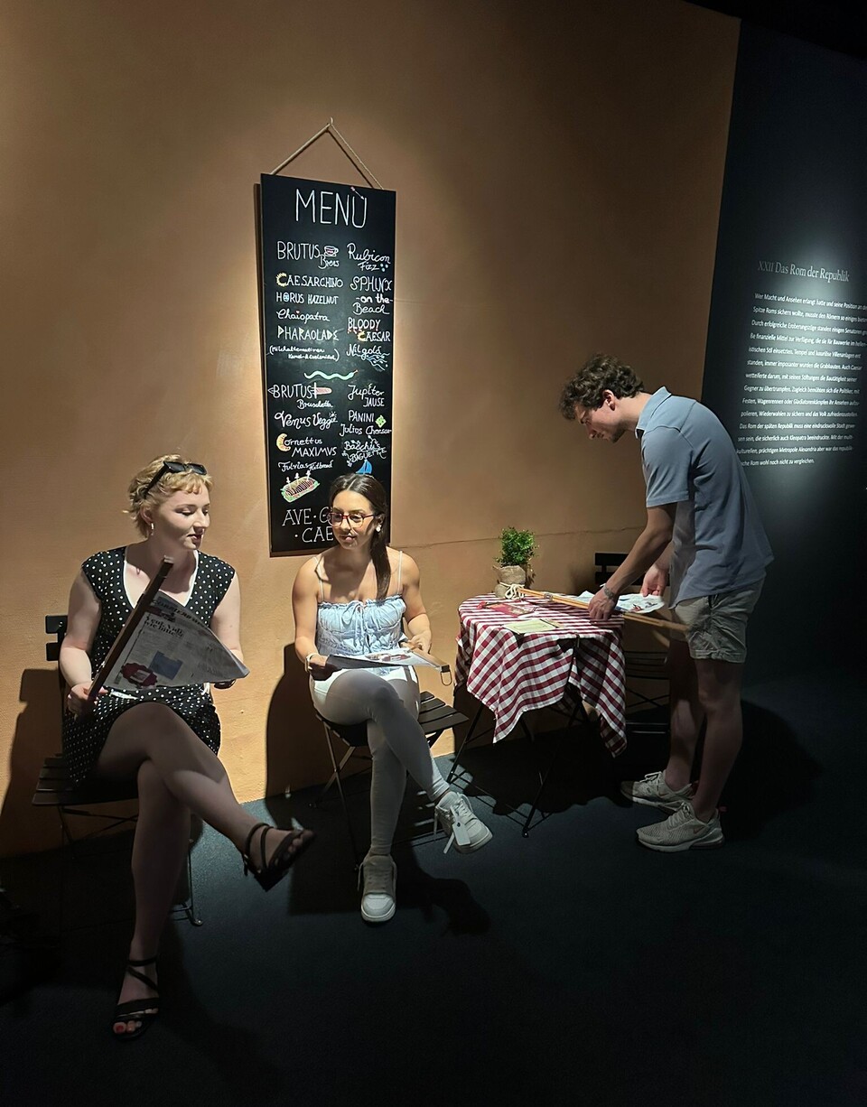 Zwei weiblich gesehene Studierende sitzen in einem italienischen Café, ein männlich gesehener Studierender steht an einem runden Bistrotisch.