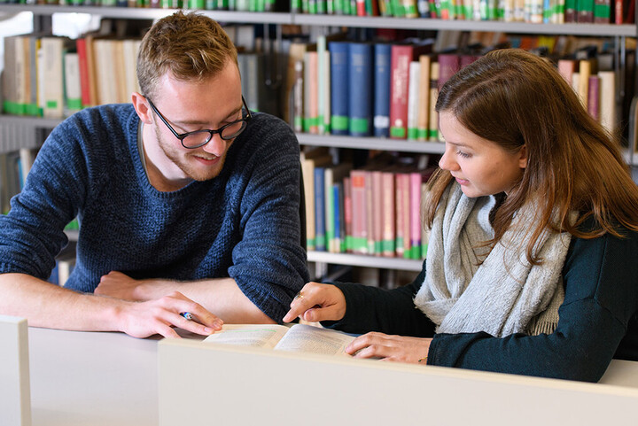 Eine Studentin zeigt einem Studenten etwas in einem aufgeschlagenen Buch.