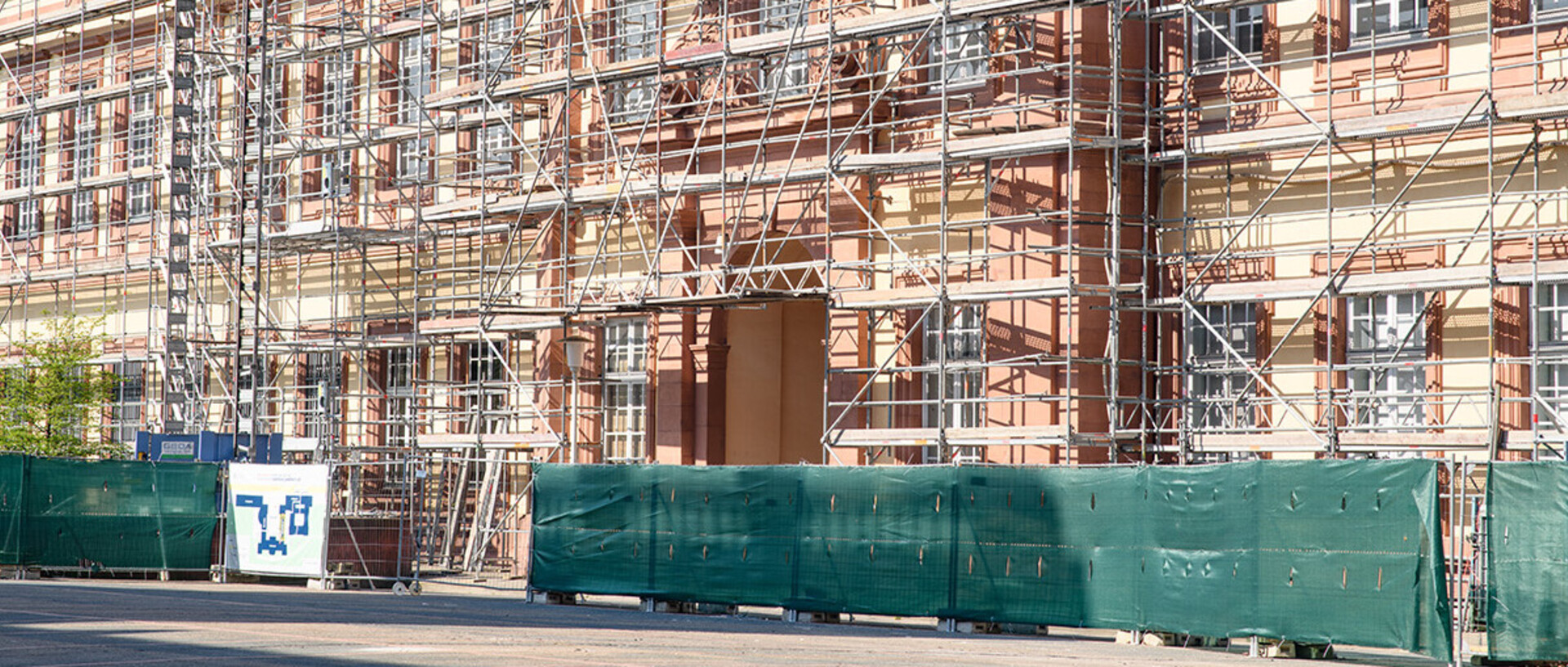 Foto der Fassade der Rückseite des Ehrenhofs Ost, die mit Baugerüsten versehen ist. Vor dem Gebäude ist eine Absperrung mit grünen Planen zu sehen. Ein Informationsbanner steht im Vordergrund auf der linken Seite.