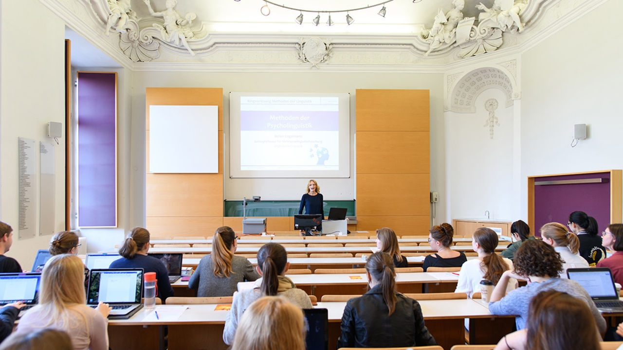 Studierende sitzen in einem hellen Hörsaal. Eine Dozentin steht vorne und hält einen Vortrag.