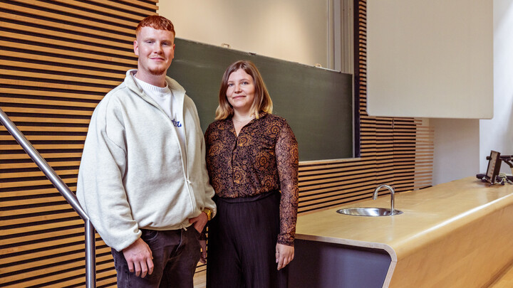 Valentin Martus und Jana Steinhauer stehen in einem Vorlesungssaal der Universität neben dem Pult.