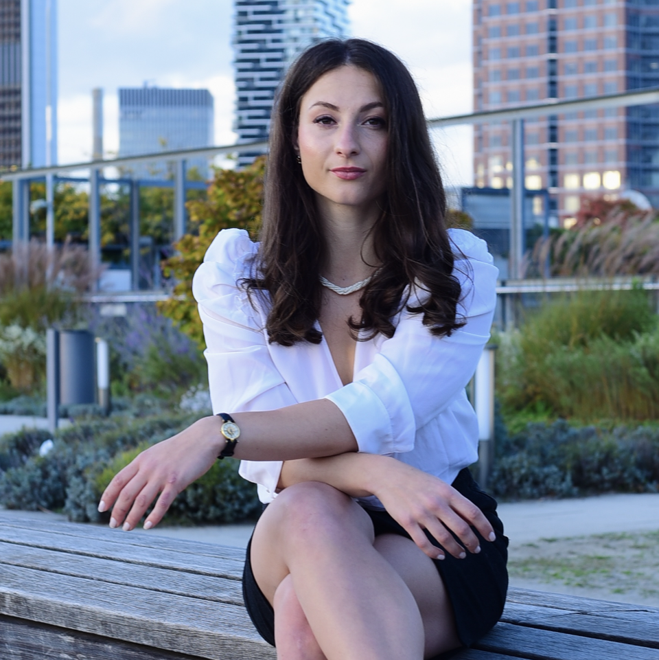 Giuliana Manganaro sitzt im Freien auf einer Bank. Sie hat lange dunkle Haare, trägt eine weiße Bluse und einen schwarzen Rock.