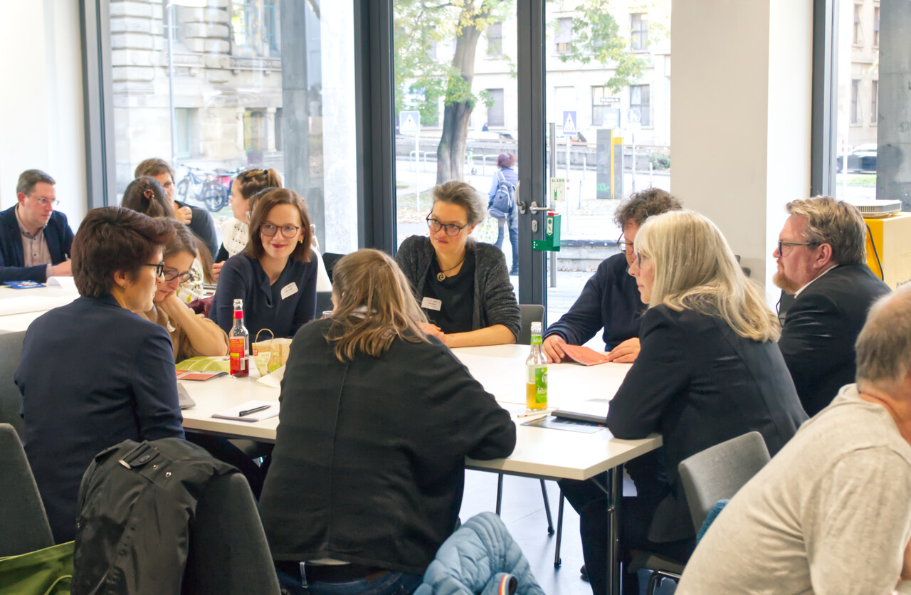 Sieben Workshop-Teilnehmende sitzen um einen Tisch herum, ins Gespräch vertieft. Der Raum blickt auf Bäume und die Stadt davor.