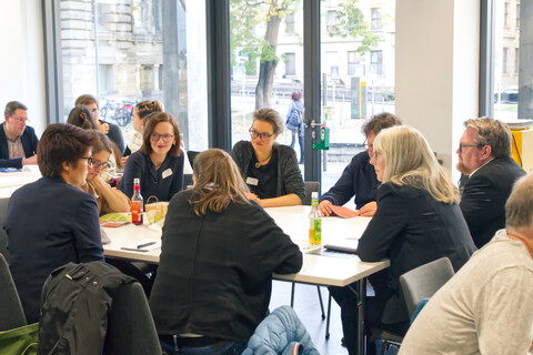 Sieben Workshop-Teilnehmende sitzen um einen Tisch herum, ins Gespräch vertieft. Der Raum blickt auf Bäume und die Stadt davor.