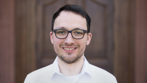 Justus Nover. He is wearing a white shirt and is standing in front of a brown door.