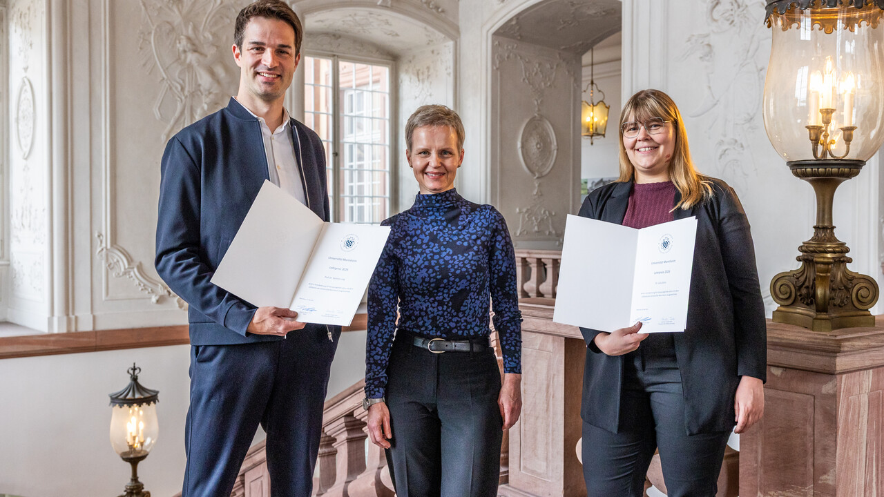 Universitätstag 2026 Drei Personen stehen nebeneinander und lächeln in die Kamera. Zwei zeigen Urkunden.