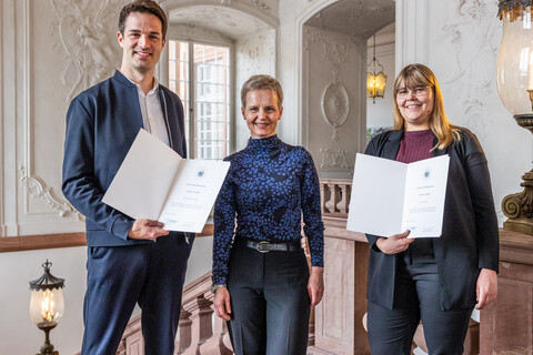 Drei Personen stehen nebeneinander und lächeln in die Kamera. Zwei zeigen Urkunden.