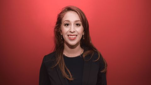 Eine lächelnde Person trägt ein schwarzes Top sowie einen schwarzen Blazer und steht vor einem roten Hintergrund. Die Person heißt Maya Moritz.